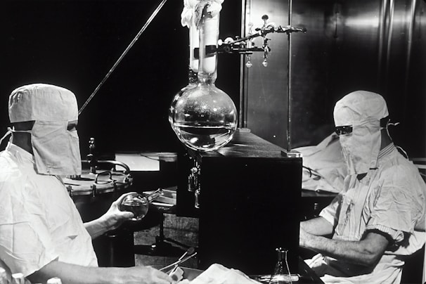 Two individuals dressed in protective clothing and masks are working in a laboratory setting. They are seated on either side of a large distillation apparatus, which includes various glassware such as a round-bottom flask. The environment suggests a high level of precision and attention to cleanliness and safety.