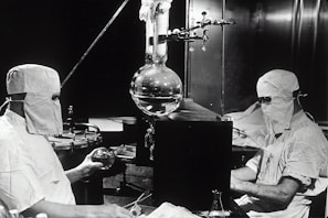 Two individuals dressed in protective clothing and masks are working in a laboratory setting. They are seated on either side of a large distillation apparatus, which includes various glassware such as a round-bottom flask. The environment suggests a high level of precision and attention to cleanliness and safety.