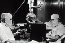 Two individuals dressed in protective clothing and masks are working in a laboratory setting. They are seated on either side of a large distillation apparatus, which includes various glassware such as a round-bottom flask. The environment suggests a high level of precision and attention to cleanliness and safety.