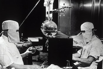 Two individuals dressed in protective clothing and masks are working in a laboratory setting. They are seated on either side of a large distillation apparatus, which includes various glassware such as a round-bottom flask. The environment suggests a high level of precision and attention to cleanliness and safety.