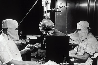 Two individuals dressed in protective clothing and masks are working in a laboratory setting. They are seated on either side of a large distillation apparatus, which includes various glassware such as a round-bottom flask. The environment suggests a high level of precision and attention to cleanliness and safety.