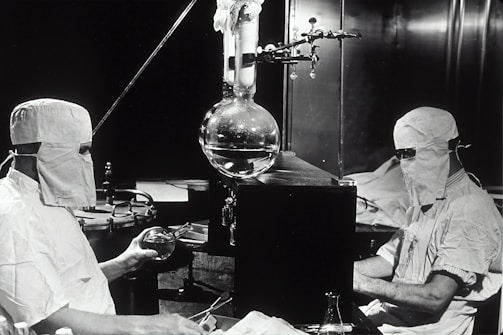 Two individuals dressed in protective clothing and masks are working in a laboratory setting. They are seated on either side of a large distillation apparatus, which includes various glassware such as a round-bottom flask. The environment suggests a high level of precision and attention to cleanliness and safety.