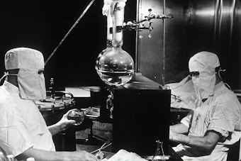 Two individuals dressed in protective clothing and masks are working in a laboratory setting. They are seated on either side of a large distillation apparatus, which includes various glassware such as a round-bottom flask. The environment suggests a high level of precision and attention to cleanliness and safety.
