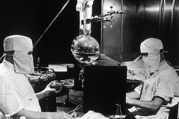 Two individuals dressed in protective clothing and masks are working in a laboratory setting. They are seated on either side of a large distillation apparatus, which includes various glassware such as a round-bottom flask. The environment suggests a high level of precision and attention to cleanliness and safety.