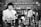 Two individuals are in a laboratory setting, surrounded by various laboratory equipment and chemical bottles. One person is working with a balance scale, while the other appears to be sitting or leaning on a bench in the background. Shelves filled with glass bottles line the wall behind them.