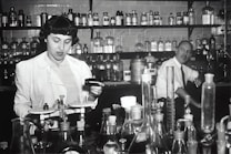 Two individuals are in a laboratory setting, surrounded by various laboratory equipment and chemical bottles. One person is working with a balance scale, while the other appears to be sitting or leaning on a bench in the background. Shelves filled with glass bottles line the wall behind them.