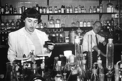 Two individuals are in a laboratory setting, surrounded by various laboratory equipment and chemical bottles. One person is working with a balance scale, while the other appears to be sitting or leaning on a bench in the background. Shelves filled with glass bottles line the wall behind them.