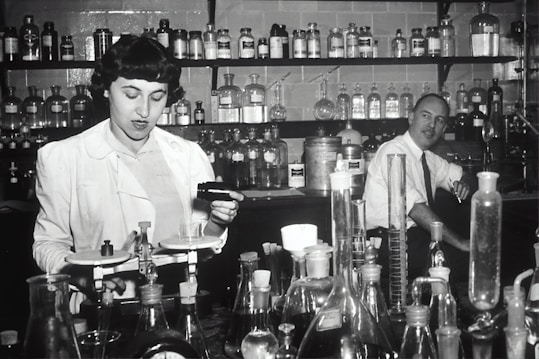Two individuals are in a laboratory setting, surrounded by various laboratory equipment and chemical bottles. One person is working with a balance scale, while the other appears to be sitting or leaning on a bench in the background. Shelves filled with glass bottles line the wall behind them.