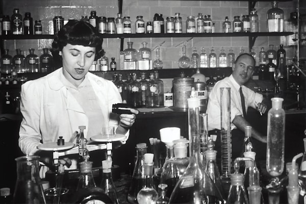 Two individuals are in a laboratory setting, surrounded by various laboratory equipment and chemical bottles. One person is working with a balance scale, while the other appears to be sitting or leaning on a bench in the background. Shelves filled with glass bottles line the wall behind them.
