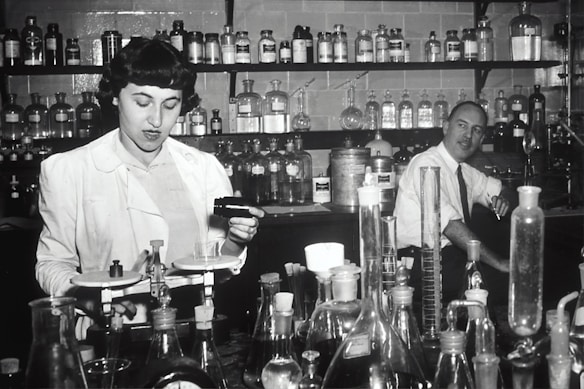 Two individuals are in a laboratory setting, surrounded by various laboratory equipment and chemical bottles. One person is working with a balance scale, while the other appears to be sitting or leaning on a bench in the background. Shelves filled with glass bottles line the wall behind them.
