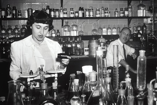 Two individuals are in a laboratory setting, surrounded by various laboratory equipment and chemical bottles. One person is working with a balance scale, while the other appears to be sitting or leaning on a bench in the background. Shelves filled with glass bottles line the wall behind them.