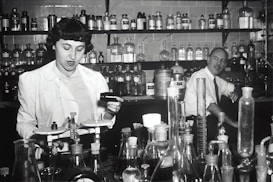 Two individuals are in a laboratory setting, surrounded by various laboratory equipment and chemical bottles. One person is working with a balance scale, while the other appears to be sitting or leaning on a bench in the background. Shelves filled with glass bottles line the wall behind them.