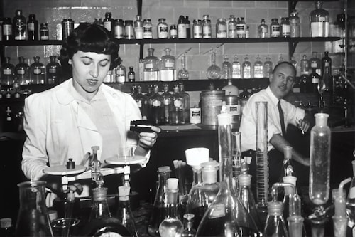 Two individuals are in a laboratory setting, surrounded by various laboratory equipment and chemical bottles. One person is working with a balance scale, while the other appears to be sitting or leaning on a bench in the background. Shelves filled with glass bottles line the wall behind them.