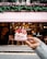 A hand holds a delicate dessert topped with whipped cream, strawberries, and decorative pink chocolate shards. The background features a stylish cafe with a pink awning and floral decorations above the entrance. The interior is softly lit, and a few people are visible sitting at tables.