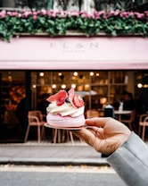 A hand holds a delicate dessert topped with whipped cream, strawberries, and decorative pink chocolate shards. The background features a stylish cafe with a pink awning and floral decorations above the entrance. The interior is softly lit, and a few people are visible sitting at tables.