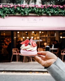 A hand holds a delicate dessert topped with whipped cream, strawberries, and decorative pink chocolate shards. The background features a stylish cafe with a pink awning and floral decorations above the entrance. The interior is softly lit, and a few people are visible sitting at tables.
