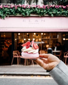 A hand holds a delicate dessert topped with whipped cream, strawberries, and decorative pink chocolate shards. The background features a stylish cafe with a pink awning and floral decorations above the entrance. The interior is softly lit, and a few people are visible sitting at tables.