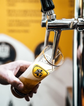 A hand is holding a tall beer glass under a tap as beer is being poured into it. The glass features a logo with a black and yellow design. The background appears to be blurred, showcasing shades of yellow and white.