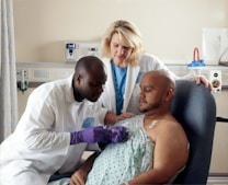 A male doctor wearing a white lab coat is attentively administering medical care to a patient. The patient, who is seated in a hospital chair, is receiving treatment through a port in his chest. A female healthcare professional stands nearby, observing and providing support.