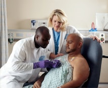 A male doctor wearing a white lab coat is attentively administering medical care to a patient. The patient, who is seated in a hospital chair, is receiving treatment through a port in his chest. A female healthcare professional stands nearby, observing and providing support.