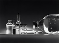 A collection of empty glass vials and containers, including ampoules and bottles, are arranged in a neat row against a dark background. The reflective surface enhances the transparency and clarity of the glassware, creating a minimalistic and clinical appearance.