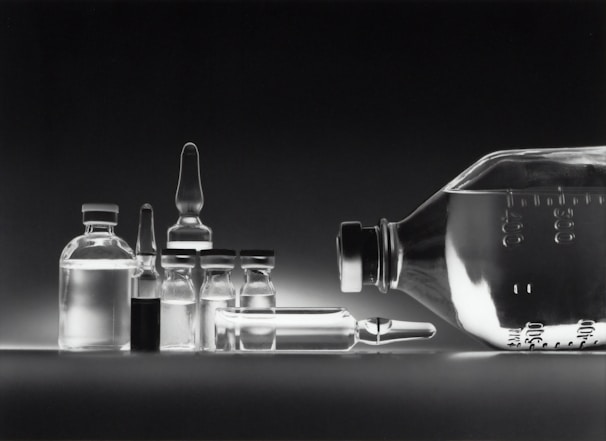 A collection of empty glass vials and containers, including ampoules and bottles, are arranged in a neat row against a dark background. The reflective surface enhances the transparency and clarity of the glassware, creating a minimalistic and clinical appearance.