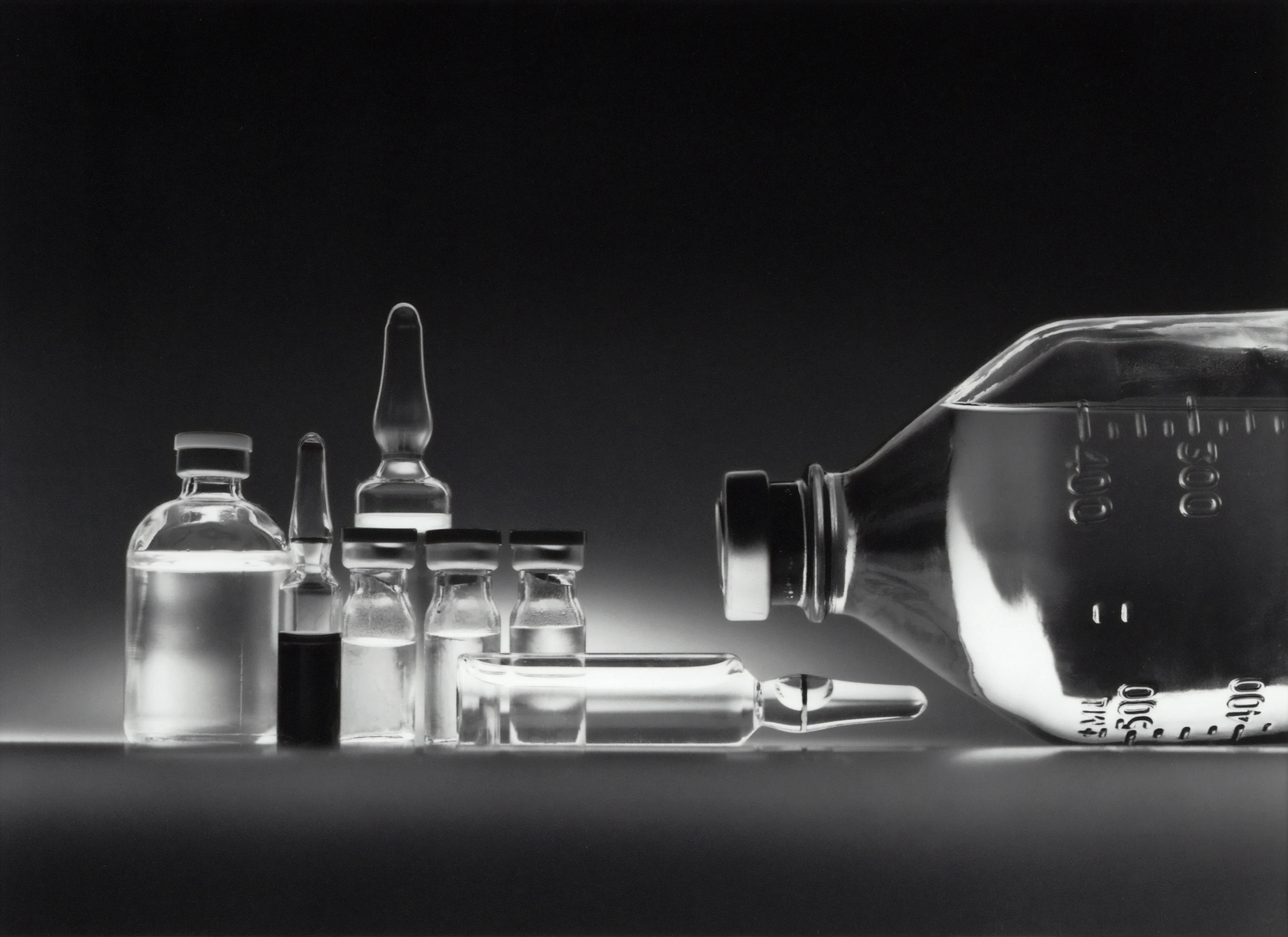 Glass medicine vials and a large infusion bottle on a dark surface, in black and white.