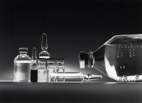 A collection of empty glass vials and containers, including ampoules and bottles, are arranged in a neat row against a dark background. The reflective surface enhances the transparency and clarity of the glassware, creating a minimalistic and clinical appearance.