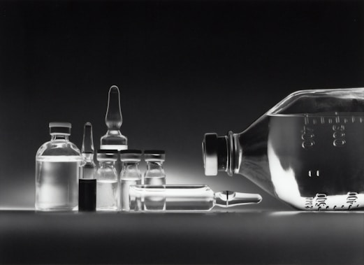 A collection of empty glass vials and containers, including ampoules and bottles, are arranged in a neat row against a dark background. The reflective surface enhances the transparency and clarity of the glassware, creating a minimalistic and clinical appearance.