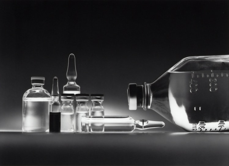 A collection of empty glass vials and containers, including ampoules and bottles, are arranged in a neat row against a dark background. The reflective surface enhances the transparency and clarity of the glassware, creating a minimalistic and clinical appearance.