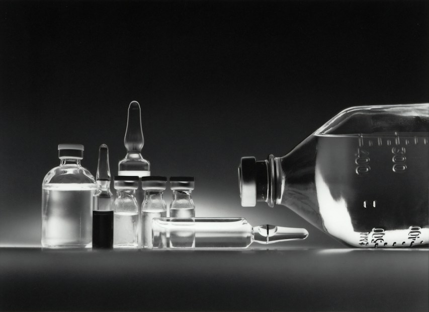 A collection of empty glass vials and containers, including ampoules and bottles, are arranged in a neat row against a dark background. The reflective surface enhances the transparency and clarity of the glassware, creating a minimalistic and clinical appearance.