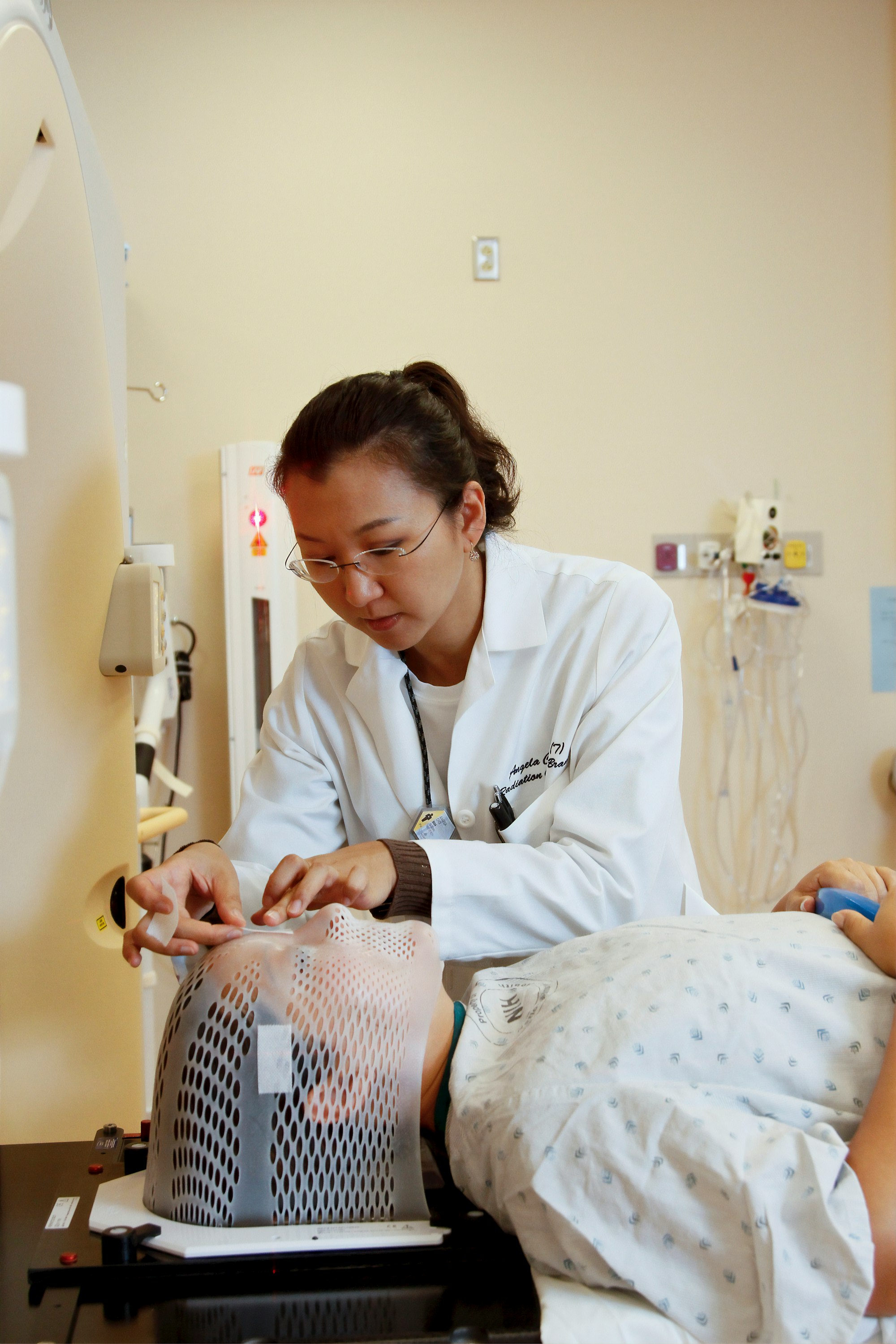 An Asian female radiation therapist places markers on a short face mask fitted to a patient model to ensure the correct position within a computed tomography (CT) scanner and later in the radiation treatment room. 2010