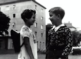 Two children face each other, smiling as they stand outside near an apartment building. One wears a patterned sweater, while the other has a traditional dress with embroidery and carries a small backpack.