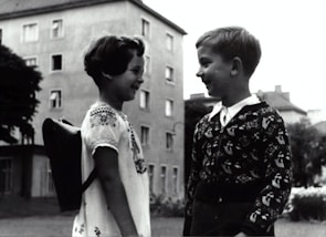 Two children face each other, smiling as they stand outside near an apartment building. One wears a patterned sweater, while the other has a traditional dress with embroidery and carries a small backpack.