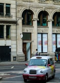 A traditional London taxi cab with a colorful design is waiting at a street intersection. The building in the background features ornate architectural details with arched windows and sculpted stonework. A few pedestrians are visible on the sidewalk.