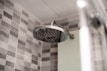A modern bathroom showerhead made of stainless steel is attached to the wall. The background features a wall tiled with gray and white checkerboard mosaic tiles, providing a clean and contemporary look.