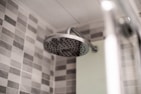 A modern bathroom showerhead made of stainless steel is attached to the wall. The background features a wall tiled with gray and white checkerboard mosaic tiles, providing a clean and contemporary look.