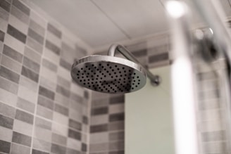 A modern bathroom showerhead made of stainless steel is attached to the wall. The background features a wall tiled with gray and white checkerboard mosaic tiles, providing a clean and contemporary look.