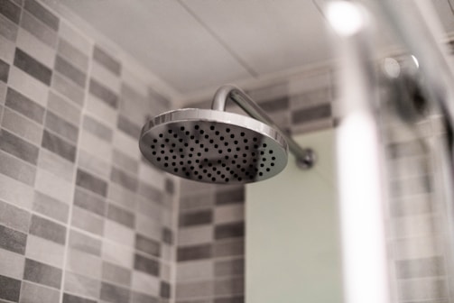 A modern bathroom showerhead made of stainless steel is attached to the wall. The background features a wall tiled with gray and white checkerboard mosaic tiles, providing a clean and contemporary look.
