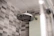 A modern bathroom showerhead made of stainless steel is attached to the wall. The background features a wall tiled with gray and white checkerboard mosaic tiles, providing a clean and contemporary look.