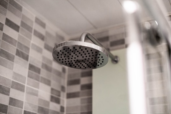 A modern bathroom showerhead made of stainless steel is attached to the wall. The background features a wall tiled with gray and white checkerboard mosaic tiles, providing a clean and contemporary look.