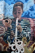 A mural depicting a young person playing a recorder. The background features swirling patterns and abstract forms, with clouds and smoke-like shapes surrounding the figure. The artwork incorporates vibrant colors and dynamic lines.