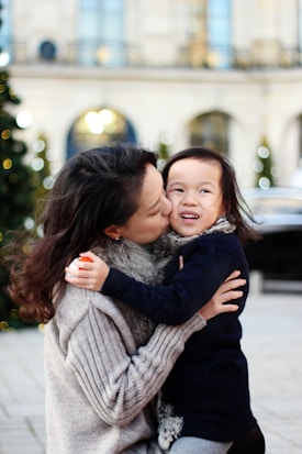 A woman wearing a cozy sweater is holding a young child close, giving them a loving kiss on the cheek. The child looks happy and is dressed warmly while they are outdoors. The background features blurred lights and a large building, adding a festive atmosphere.