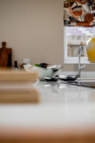 A modern kitchen area with a white countertop featuring a decorative bowl containing fruits. In the background, a wooden cutting board is positioned vertically, and there's a bottle next to it. A colorful window shade with floral patterns partially covers a window above a silver faucet. The overall look is minimalistic and stylish.