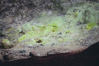 A rocky surface with distinct greenish-yellow mineral deposits scattered across the area. The terrain appears to be uneven and rugged, showing signs of erosion and mineral accumulation.