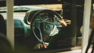 A view through a vehicle window showing a person gripping a steering wheel. The interior of the car is visible, including the dashboard and speedometer. The lighting is warm, creating a soft reflection on the glass.