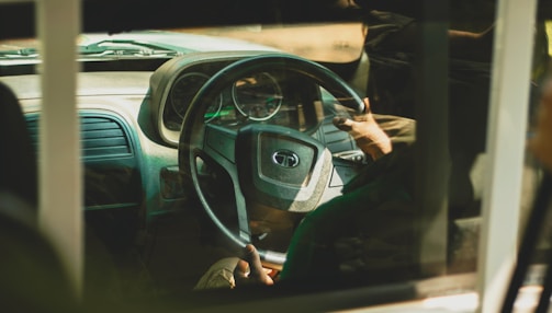 A view through a vehicle window showing a person gripping a steering wheel. The interior of the car is visible, including the dashboard and speedometer. The lighting is warm, creating a soft reflection on the glass.