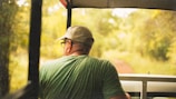 A person wearing a green shirt and a beige cap is seated in an open vehicle, looking out at a lush, blurred green forest. The image is taken from behind the person, capturing the serene natural surroundings and the sense of travel or exploration.