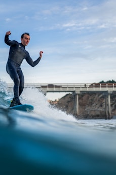 A surfer dressed in a black wetsuit rides a wave with a confident stance. The background features a long pier jutting out from the shore and rugged cliffs framed against a partially cloudy sky.