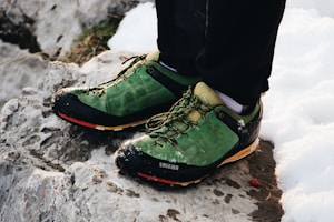 A pair of green hiking shoes with black and yellow accents are worn by someone standing on rocky terrain next to snow. The shoes appear slightly muddy and have a rugged design suitable for outdoor activities.